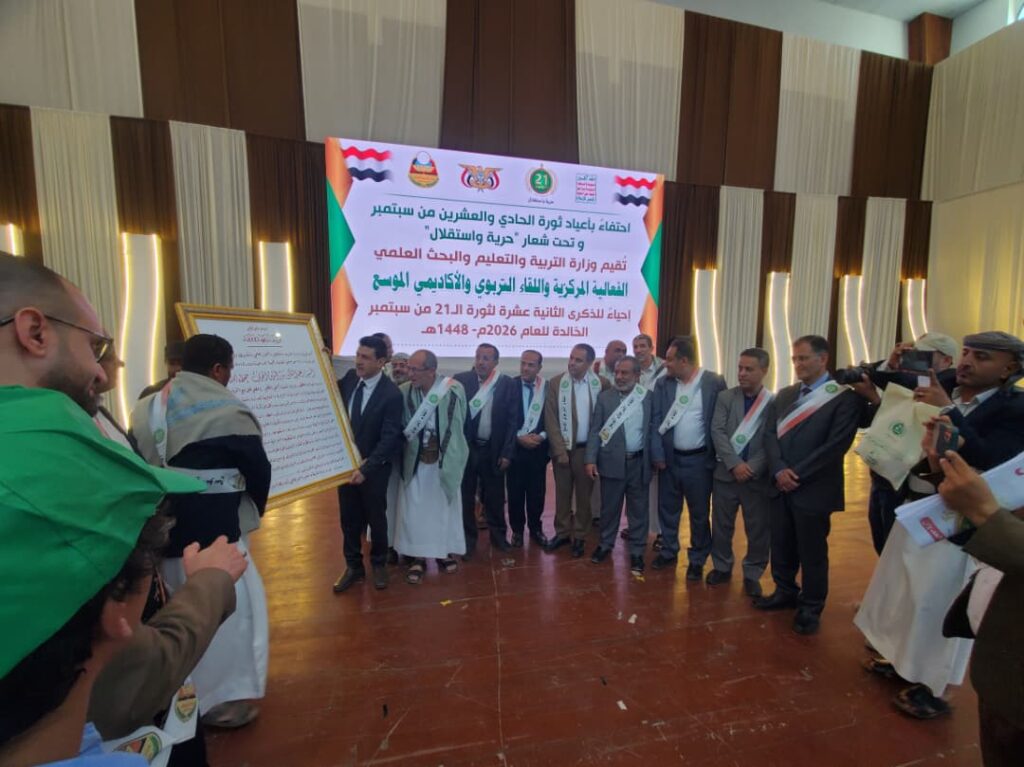 IMG-20260919-WA0055 - جامعة 21 سبتمبر Group of men in suits and traditional dress pose during a formal ceremony in a hall, with a framed certificate visible on the left.