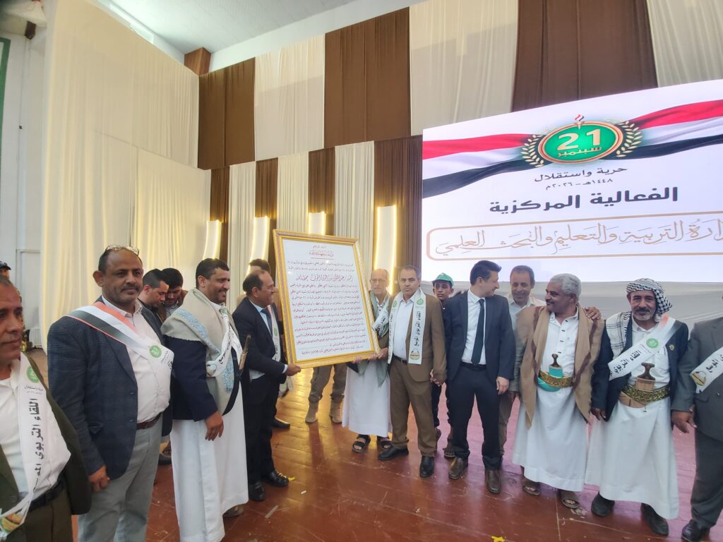 IMG-20260919-WA0058 - جامعة 21 سبتمبر Group of men in traditional robes and suits at an indoor award ceremony, some wearing sashes, holding a large framed document against a backdrop with a banner featuring Arabic text and a 21-year emblem.