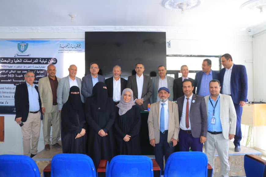 Group of professionals posing for a photo at a formal event, with a banner on the left and a large screen behind them in a conference room.