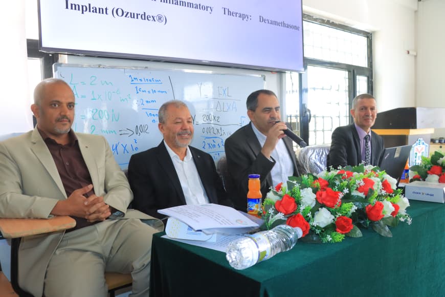 Four men at a conference panel; one speaks into a microphone, with flowers on the table and a whiteboard behind.