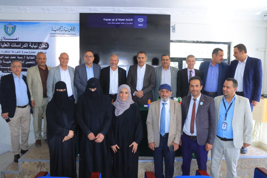 Group of men in suits and women in black abayas posing for a photo during a formal ceremony or event. In the background, a banner and large screen suggest a conference setting.