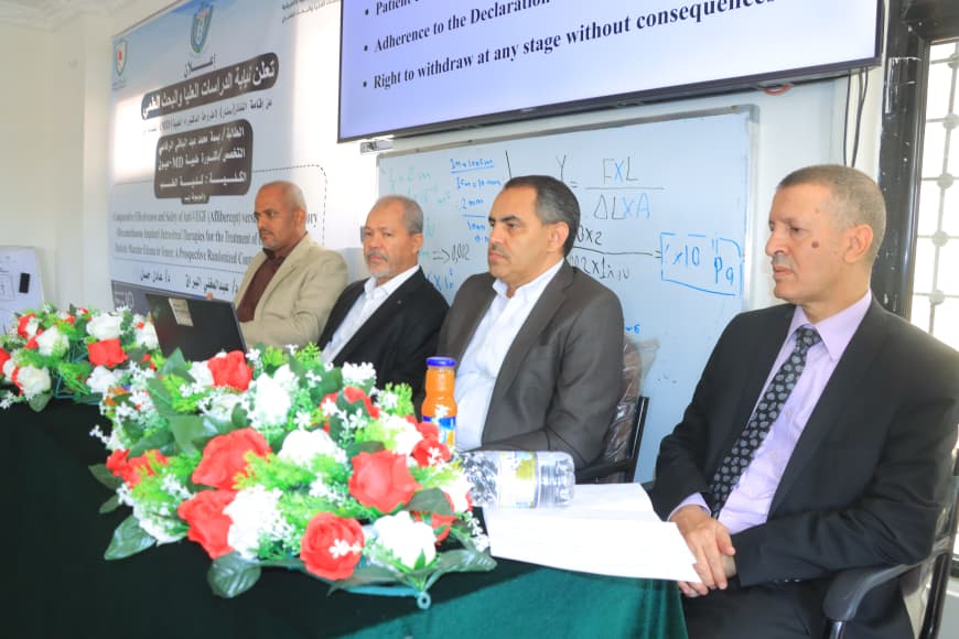 Panel of four men in suits seated at a conference table with a flower centerpiece, presenting to an audience in a classroom or lecture hall.