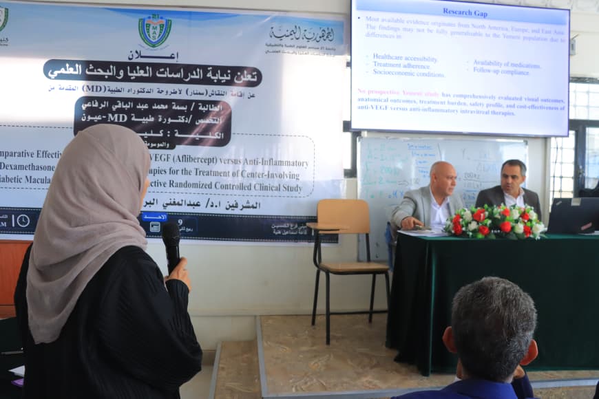 Female speaker in a hijab addresses a panel at a medical conference; a banner in Arabic and two men seated at a table with flowers behind.