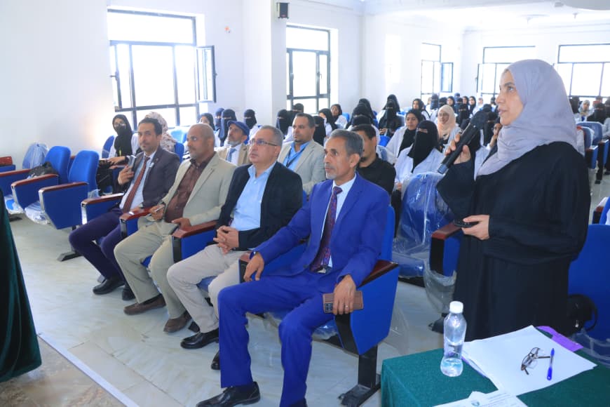 Woman wearing a hijab speaks into a microphone to a seated conference audience in a bright room.