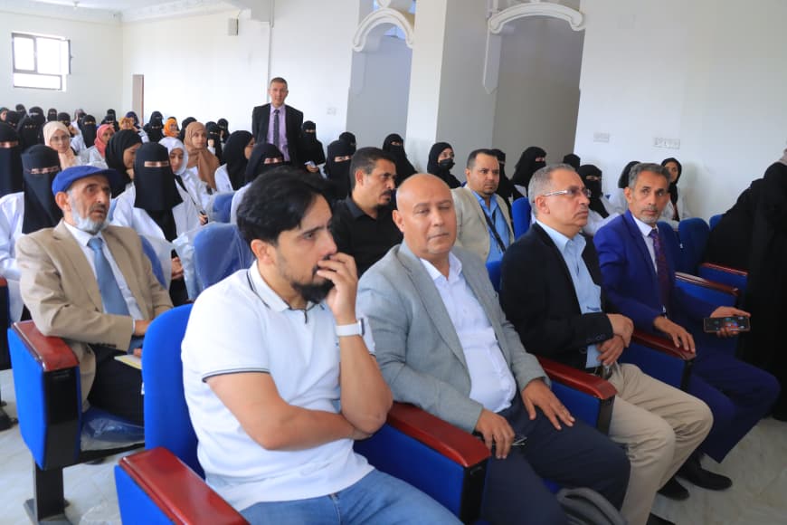 Audience seated in a bright hall for a formal event; front-row men with suits, women in hijabs behind.