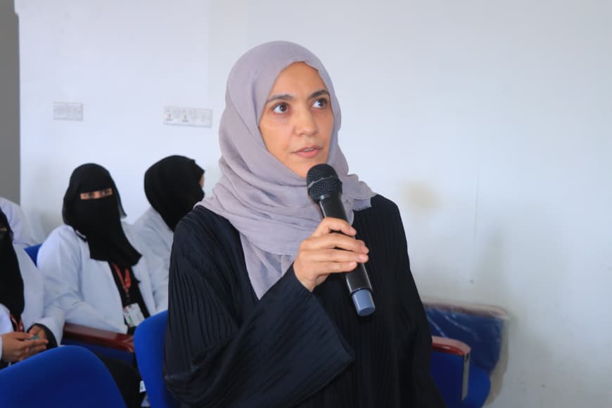 Woman wearing a gray hijab speaks into a handheld microphone in a conference room with seated attendees in the background.