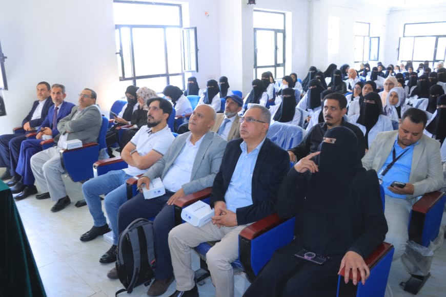Group of men and women seated in a bright conference hall, many women wearing black niqabs, listening to a presentation.