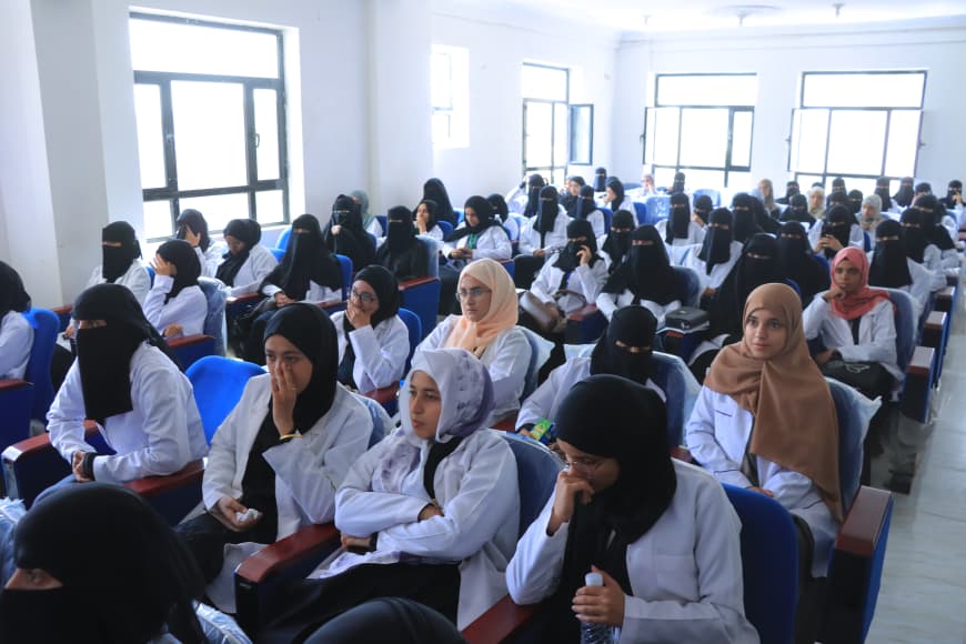 Large classroom filled with female students in white coats wearing hijabs, seated in blue chairs.