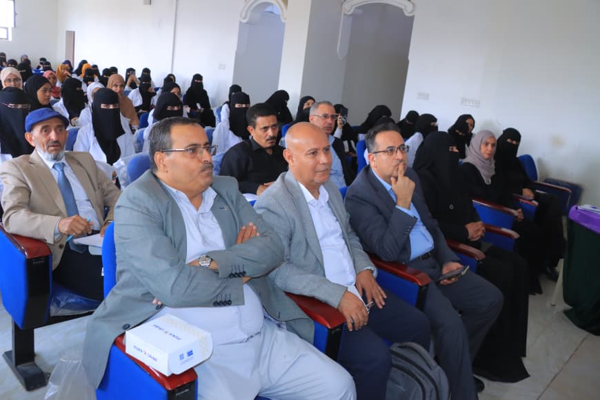 Audience seated in a conference room, front-row men in light-colored jackets and women in hijabs in the background.