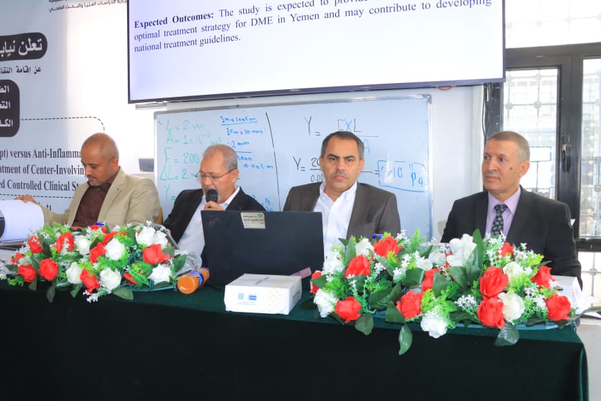 Panel of four men in suits seated at a conference table with a microphone and laptop, floral centerpiece in front.