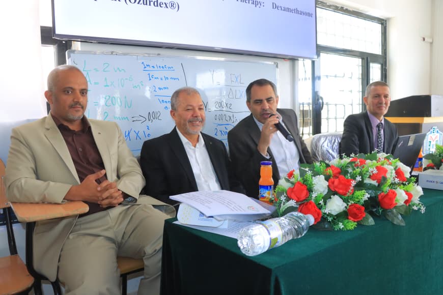 Panel of four men in suits seated at a conference table; one is speaking into a microphone, with a flower arrangement, papers, and water bottles on the table, and a whiteboard with equations behind them.
