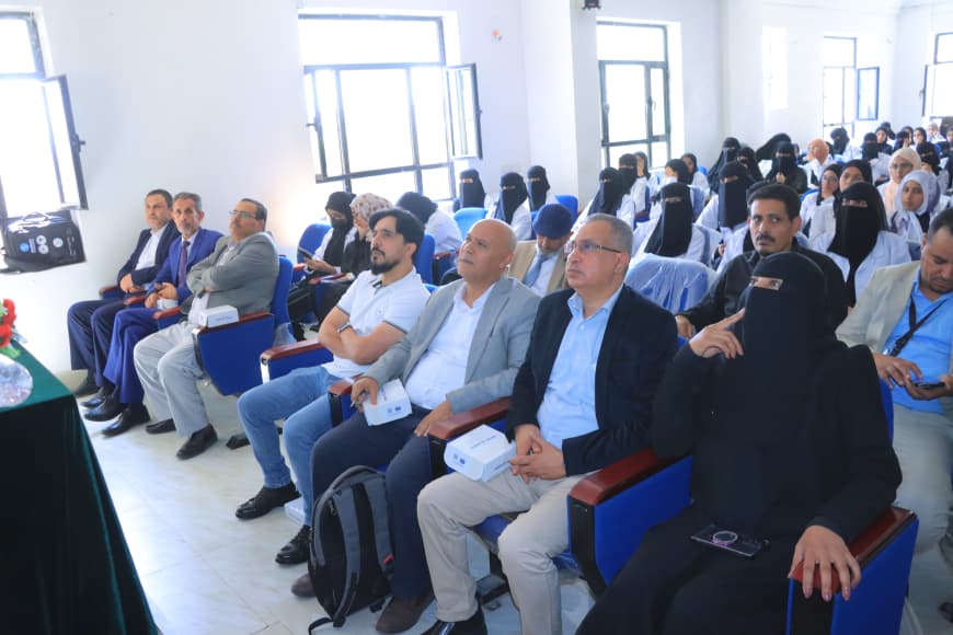 Audience seated in a bright classroom or auditorium, front-row men in suits and a woman in black niqab among attendees during a formal event.