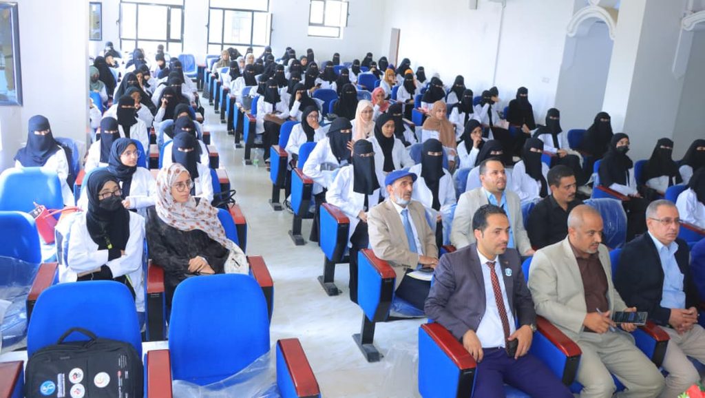 Lecture hall packed with students, many women in niqab, seated in blue chairs for a seminar or lecture.