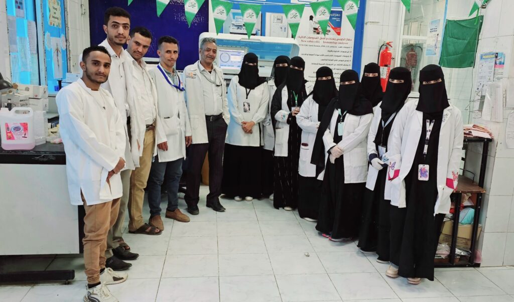 Group of medical staff posing for a photo in a lab, several women in black hijabs and white coats lined up against the wall.