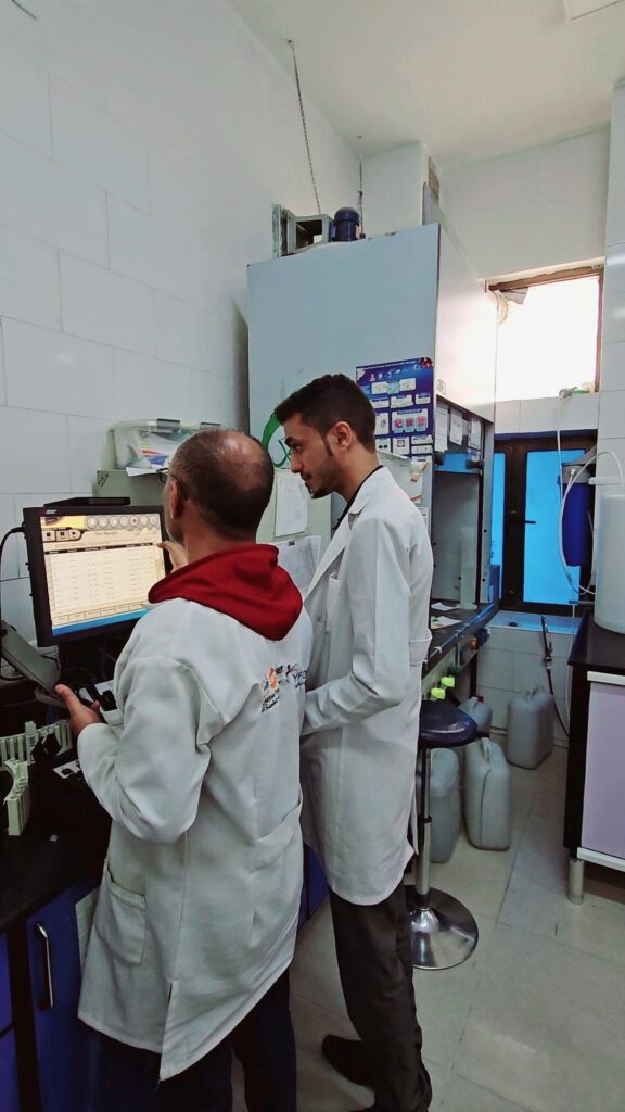 Two lab researchers in white coats stand at a bench, focusing on a computer screen displaying test data in a clean laboratory setting.