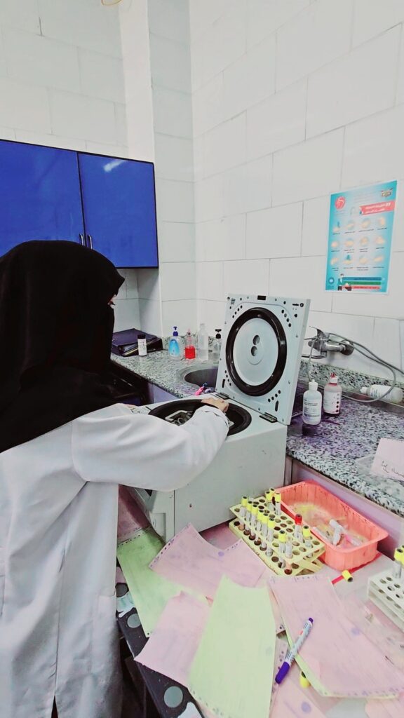 Lab technician in white coat operating a benchtop centrifuge with its lid open on a tiled lab bench, surrounded by tubes and paperwork.