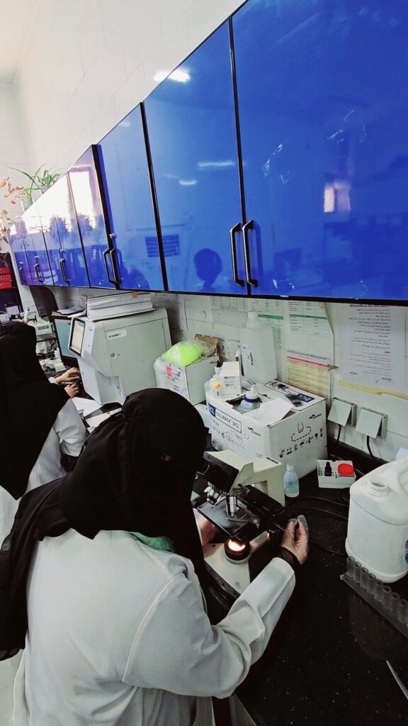 Laboratory scene with a technician in a white coat and hijab using a microscope under blue upper cabinets at a cluttered bench, shelves in view.