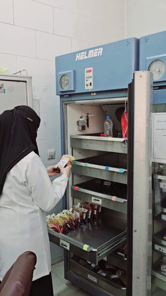 Lab worker in a white coat and hijab places a wrapped item into an open blue Helmer storage freezer with labeled shelves and drawers in a clinical setting