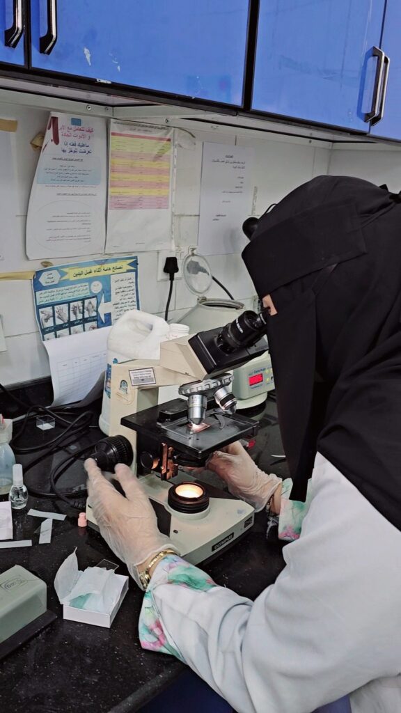 Scientist in a hijab and gloves using a compound microscope at a lab bench with papers and posters on the wall behind.