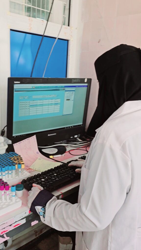 Lab technician in a white coat and hijab typing at a computer, with a row of color-coded test tubes and notes on the desk nearby.