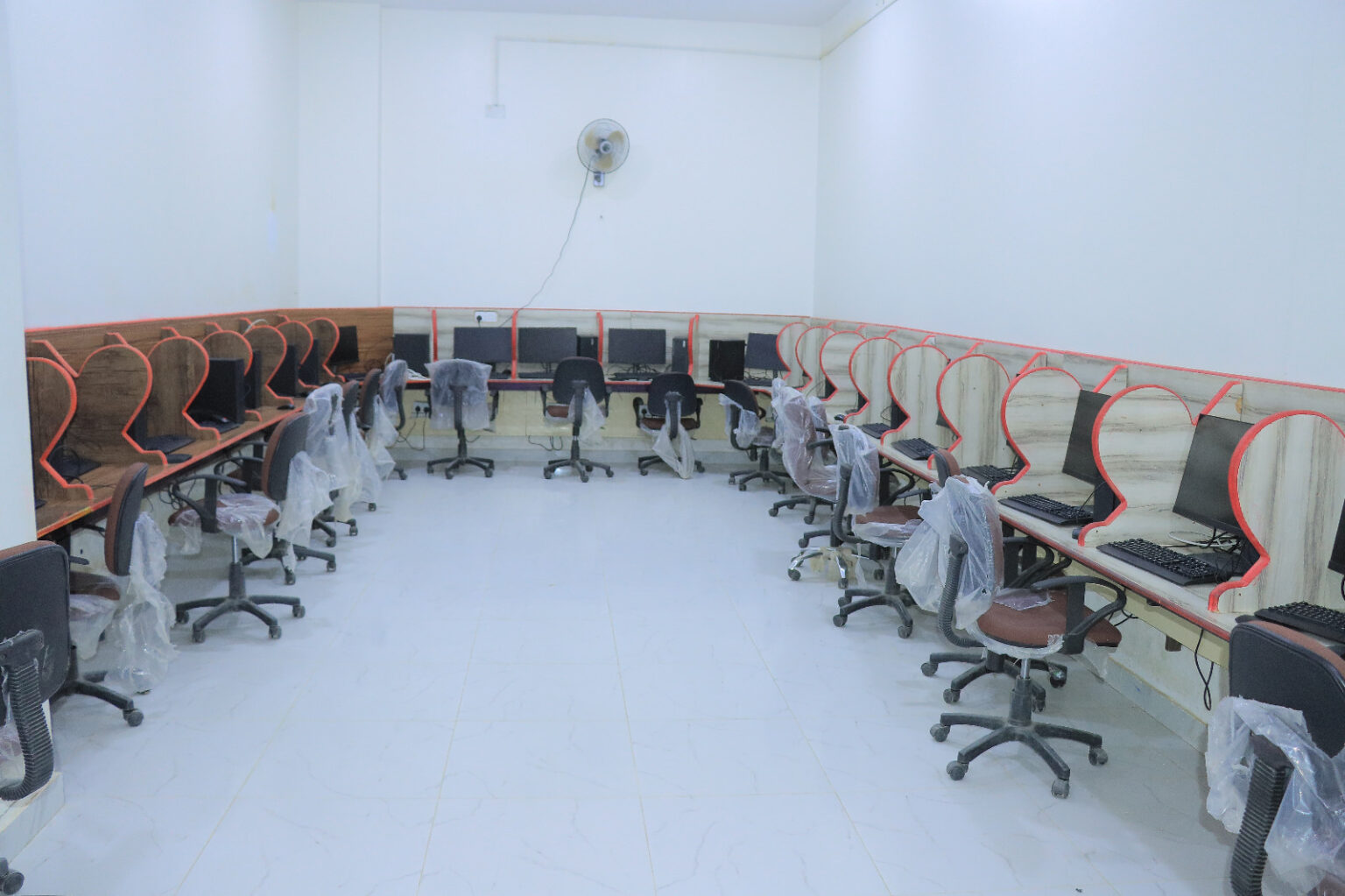 Empty computer lab with rows of desks and monitors along the walls; chairs are wrapped in plastic.