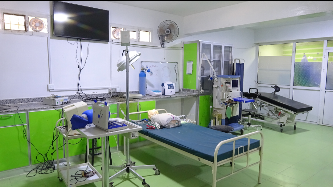 Hospital procedure room with a blue patient bed, a mannequin on the bed, overhead lamp, monitor, and green cabinets.