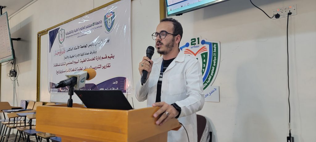 Man in a white lab coat speaks into a microphone at a wooden podium in a classroom, with a large Arabic banner behind.