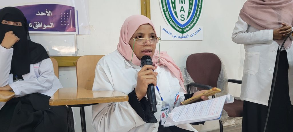 Woman in a pink hijab speaks into a handheld microphone while seated at a desk, holding a notebook and papers in a classroom setting.