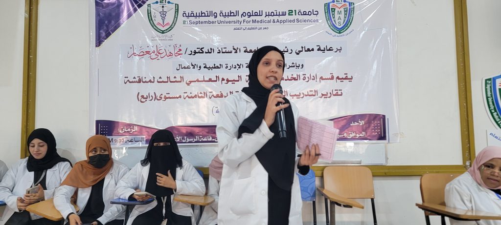 Woman in a hijab speaks into a microphone at a student panel, with peers in lab coats seated behind and a banner in Arabic banner in the background.