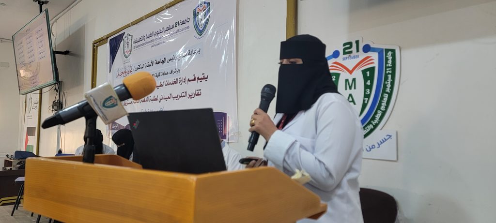 Woman wearing a niqab and white gown speaks into a microphone at a podium during a formal event, with an Arabic banner behind her.