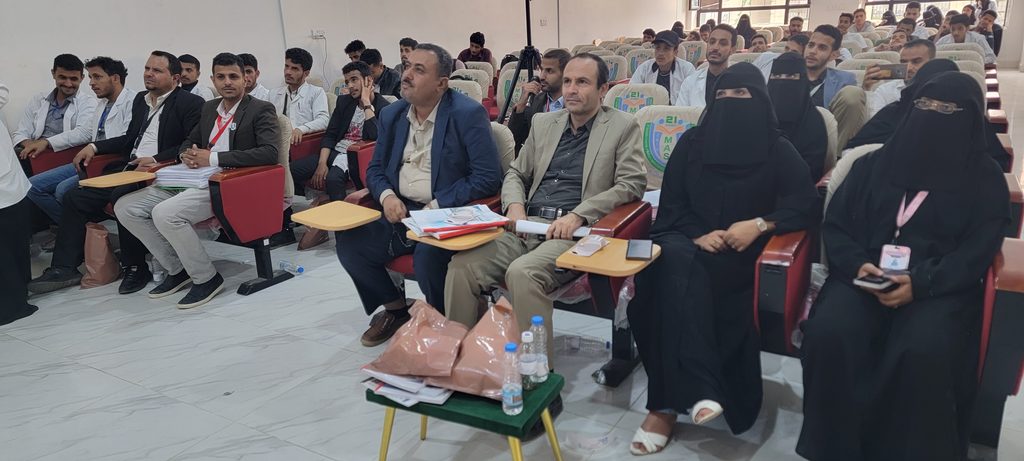 Audience in a lecture hall: men in suits and casual wear, women in black abayas, seated and listening to a presentation.
