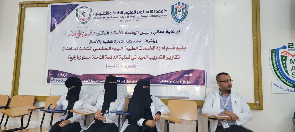 Group of medical students and a man in a white coat seated in a classroom with a large Arabic banner about a university event behind them